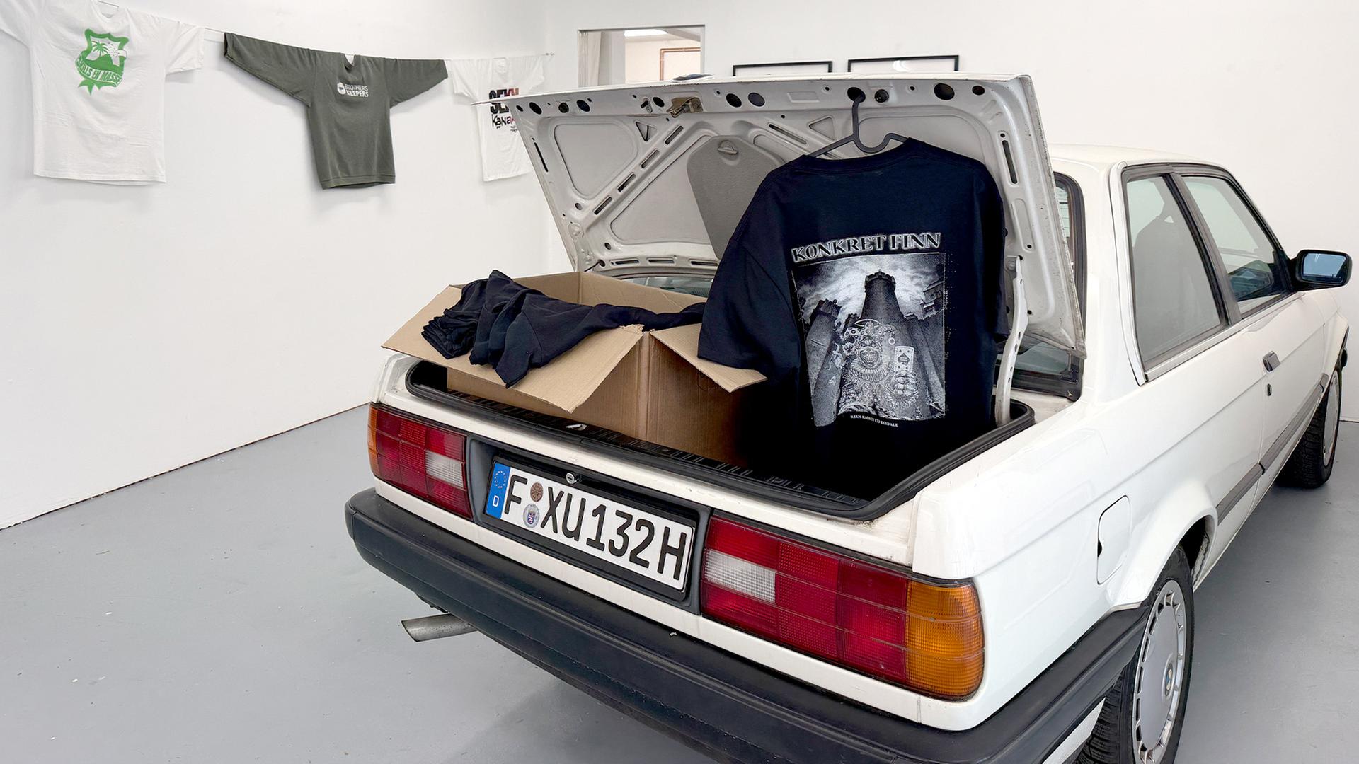 Ausstellungsraum mit einem Auto in der Mitte, dessen Kofferraum geöffnet ist und dort ein Karton und ein aufgehängtes bedrucktes T-Shirt zu sehen ist. Im Hintergrund an den Wänden weitere bedruckte Sweatshirts. | Christiane Schwalm/hr
