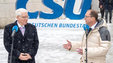 Josef Schuster und Alexander Dobrindt sprechen vor einem CSU-Schild. Es schneit.