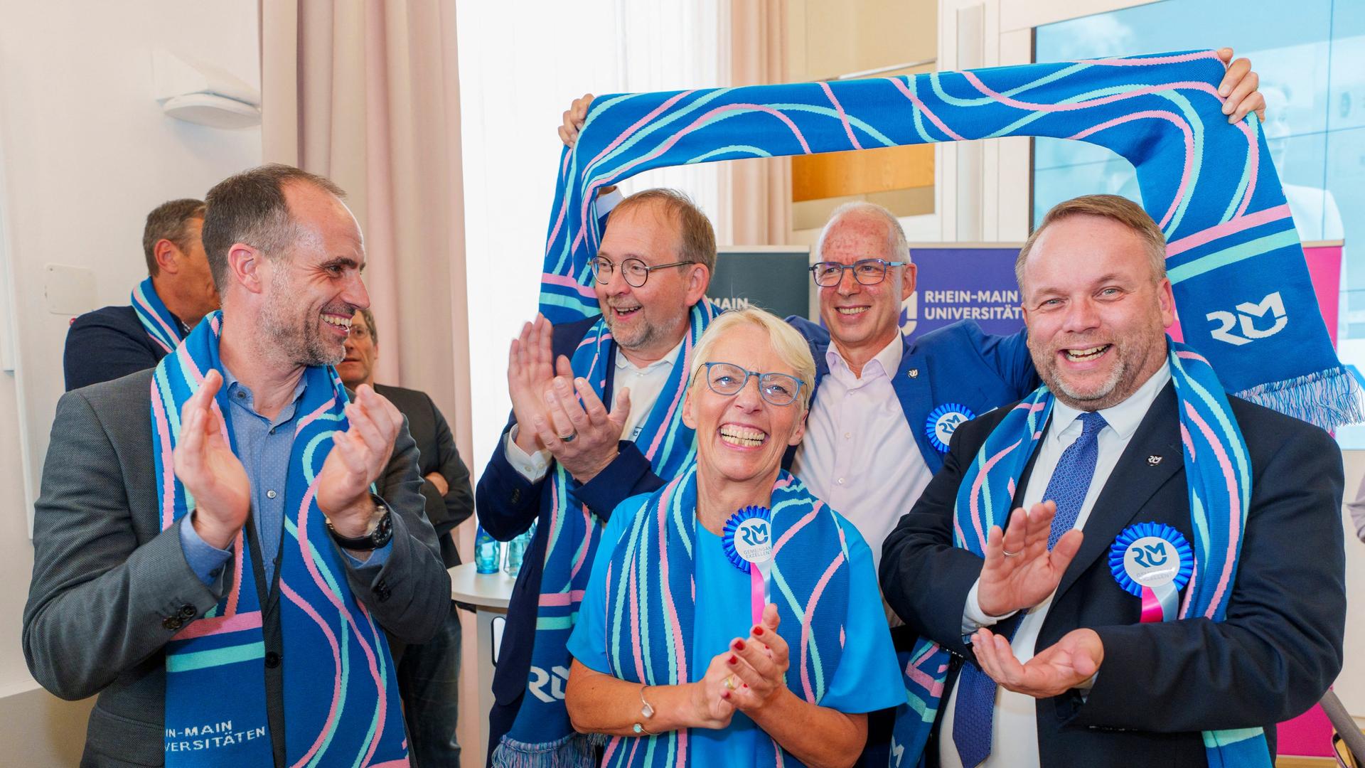 Fünf Personen mit Schals der Rhein-Main-Universitäten stehen lächelnd zusammen und feiern den Exzellenz-Titel. | picture alliance/dpa | Andreas Arnold