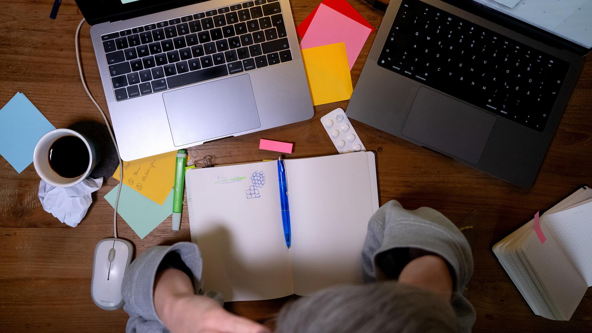Eine Person sitzt mit zwei Laptops und einem Notizbuch an einem Schreibtisch. | dpa