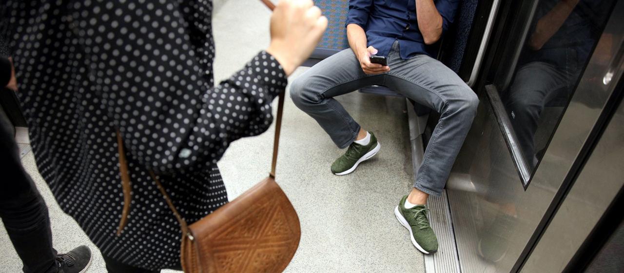Ein Mann sitzt in der U-Bahn mit gespreizten Beinen und hält ein Smartphone in der Hand. 