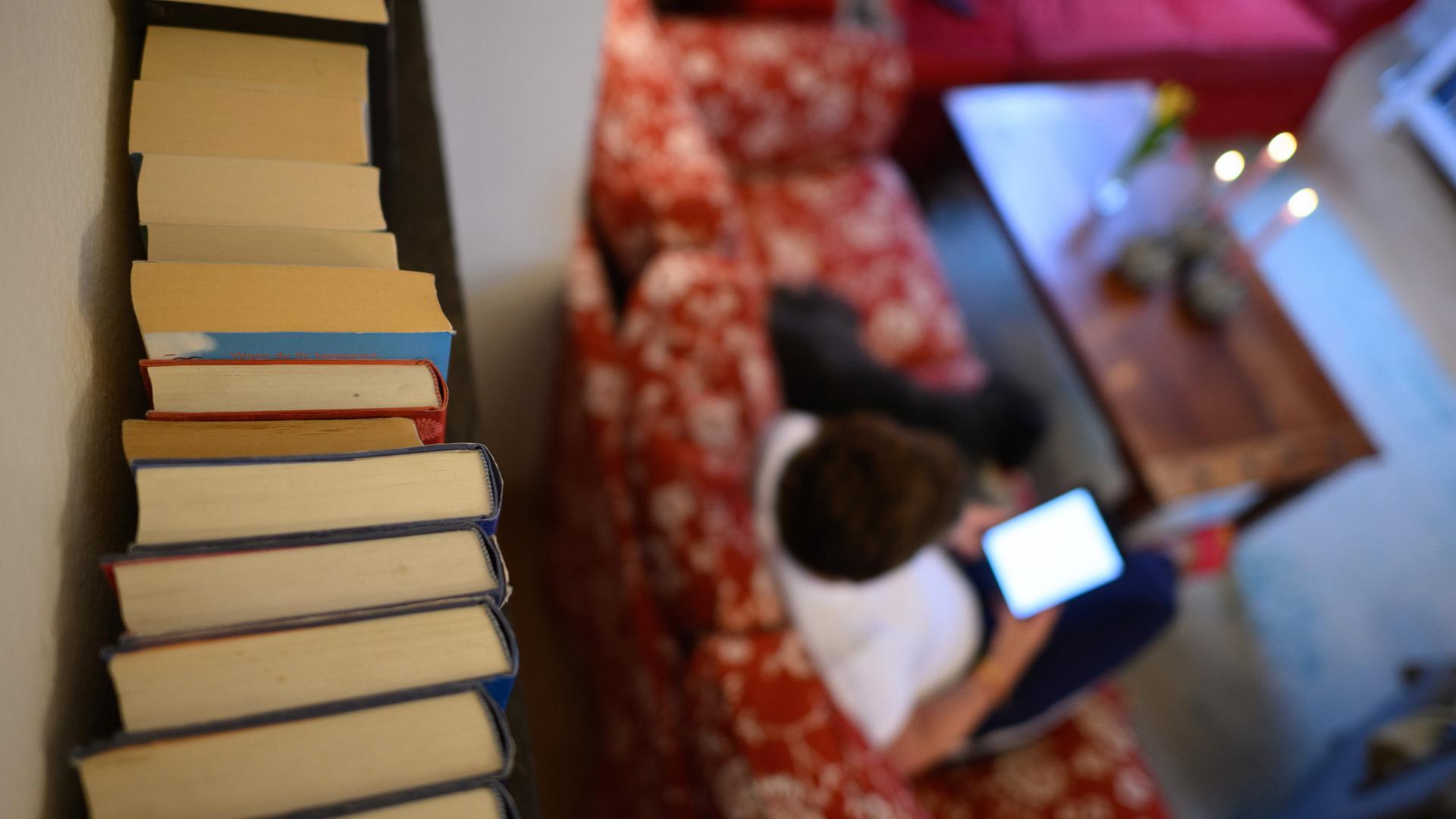 Bücher stehen in einem Wohnzimmer im Regal, während eine Frau mit einem Tablet auf dem Sofa sitzt. | picture alliance/dpa