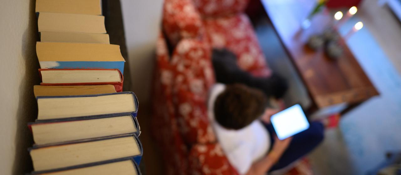 Bücher stehen in einem Wohnzimmer im Regal, während eine Frau mit einem Tablet auf dem Sofa sitzt.