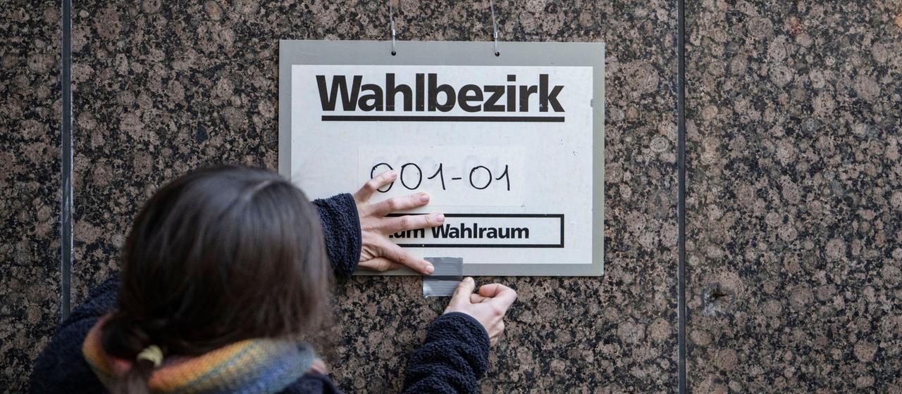 Eine Wahlhelferin befestigt ein Schild mit der Aufschrift "Wahlbezirk" an einer Wand.