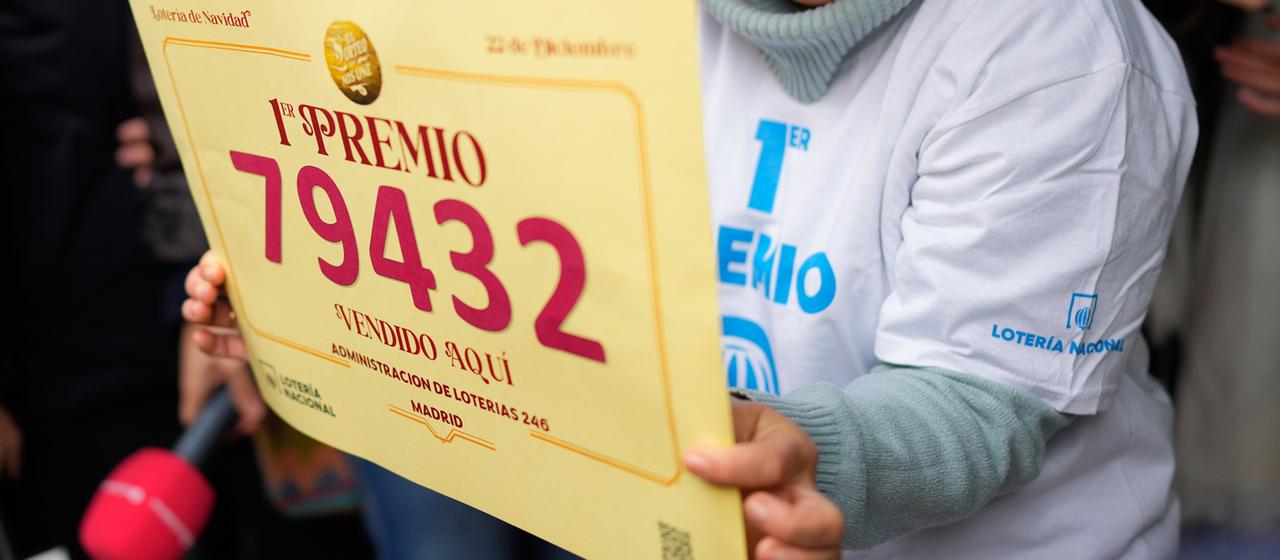 Eine Losverkäuferin hält ein Schild mit der Gewinnernummer 79.432 hoch.