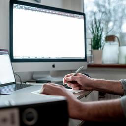 Ein Mann sitzt an einem Tisch vor einem Monitor und einem Laptop.