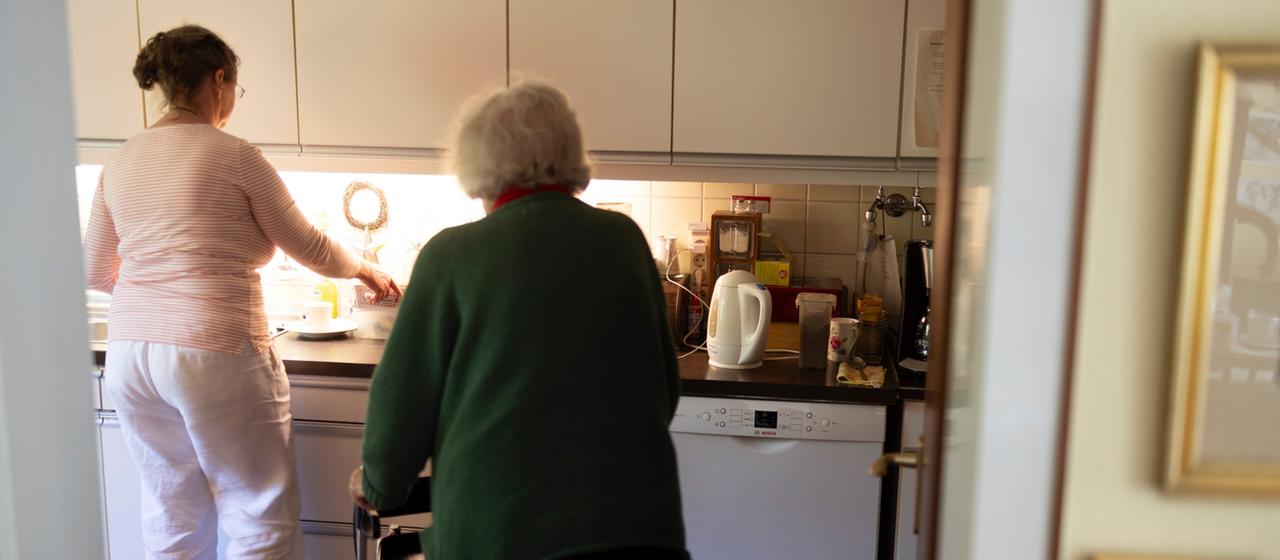 Eine jüngere Frau und eine ältere Frau mit Rollator sind in einer Küche zu sehen.