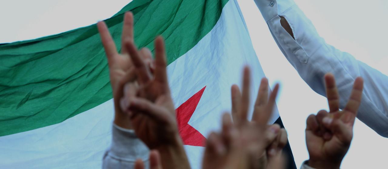 Menschen halten vor der syrischen Flagge jeweils zwei Finger in die Höhe - das Friedenszeichen.