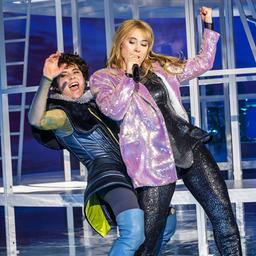 Ein blonde Frau in schwarzem Glitzeranzug steht auf einer grün erleuchteten Bühne und singt in ein Mikro.