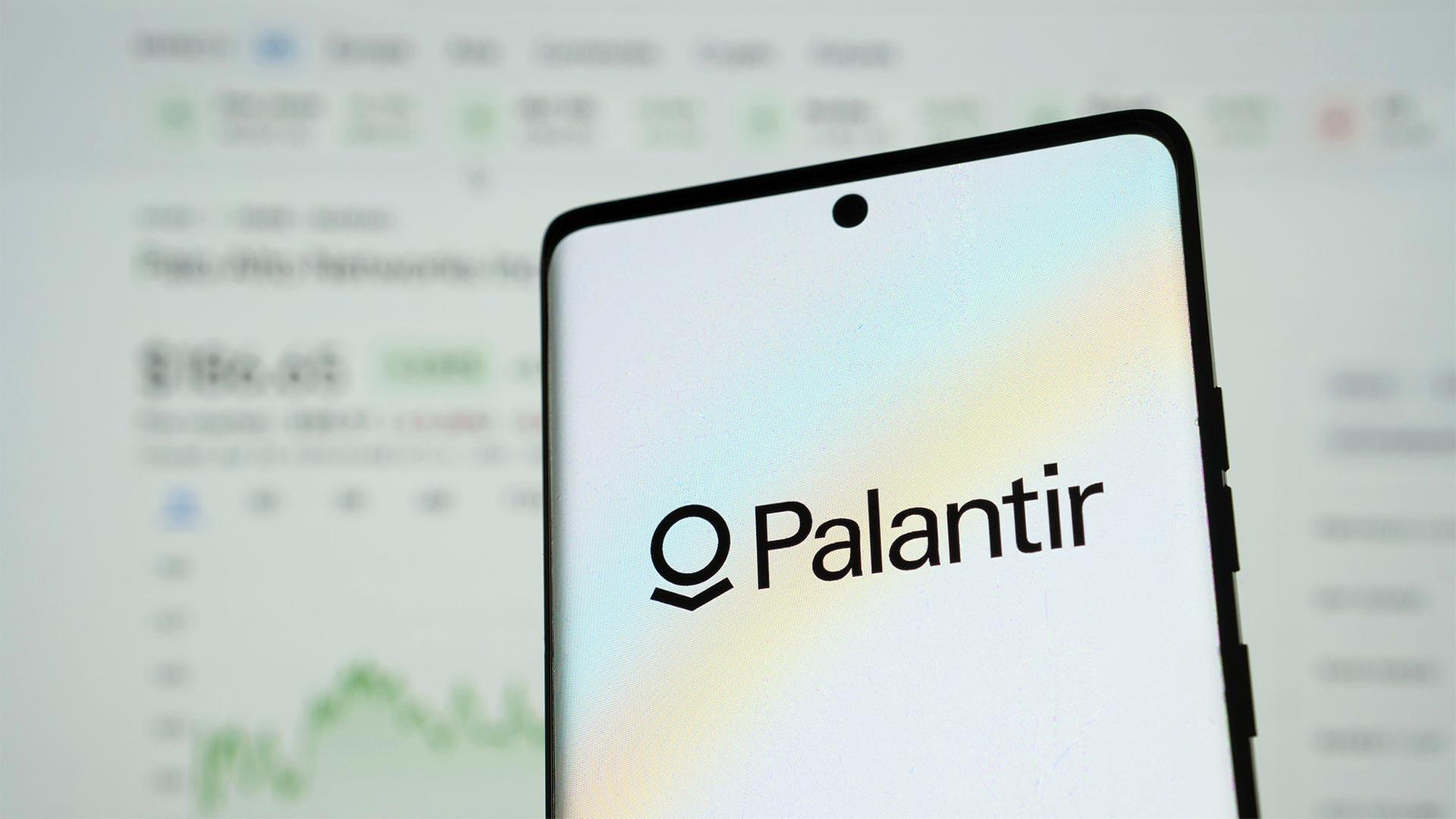 Palantir Logo auf einem Smartphone mit unscharfen Finanzdaten im Hintergrund. | picture alliance / imageBROKER