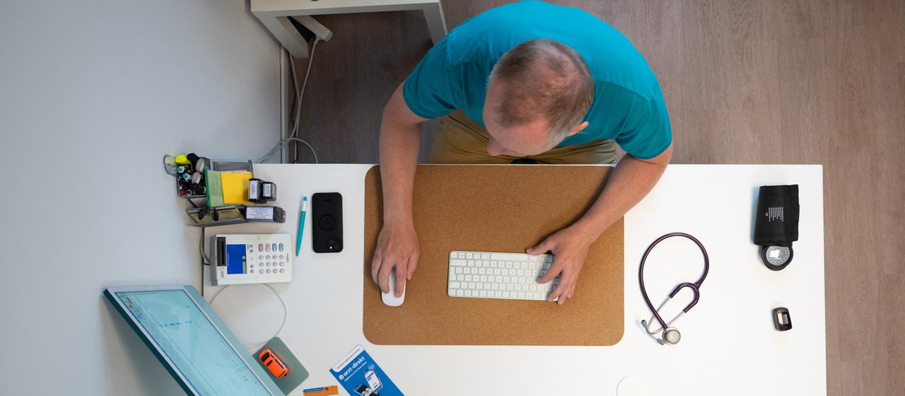 Ein Hausarzt sitzt in einem Sprechzimmer seiner Hausarztpraxis an einem Schreibtisch.
