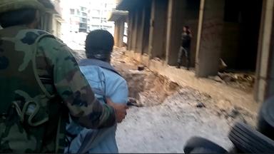 Ein Mann in Flecktarn führt einen anderen Mann zu einer Grube zwischen zwei Häusern.