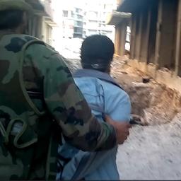 Ein Mann in Flecktarn führt einen anderen Mann zu einer Grube zwischen zwei Häusern.