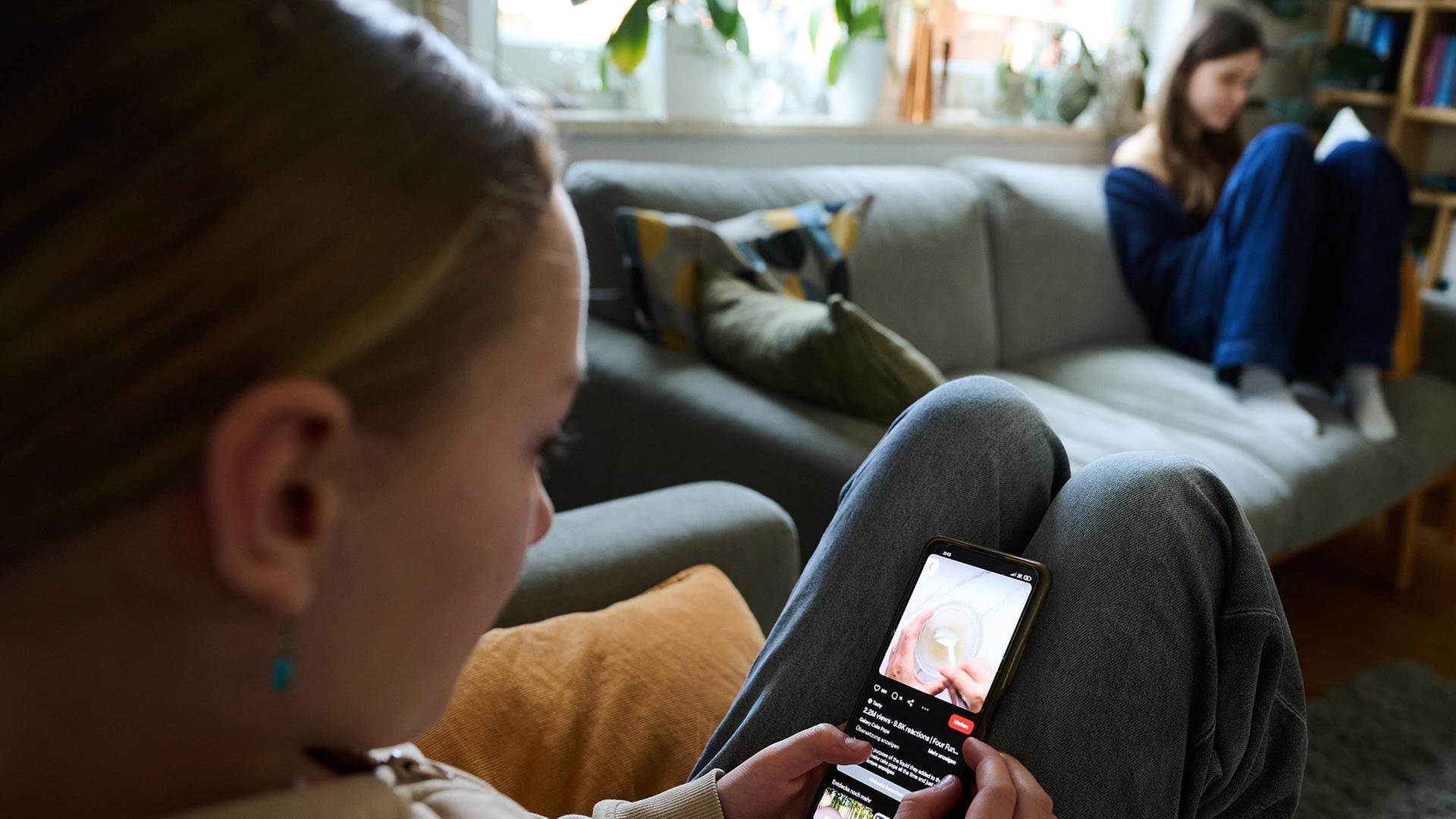 Ein Mädchen sitzt auf dem Sofa und guckt in ihr Smartphone. | picture alliance/dpa