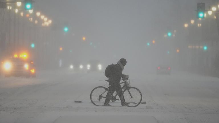 Eine Person schiebt bei starkem Schneefall ein Fahrrad über eine verschneite Straße.