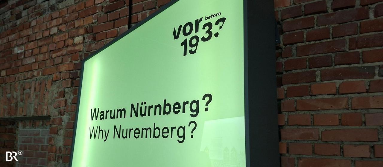 Grüne Ausstellungswand mit der Aufschrift "Warum Nürnberg?". Sie steht vor einer Wand aus roten Ziegeln. Das zeigt den unfertigen Charakter der von Hitler geplanten Kongresshalle.