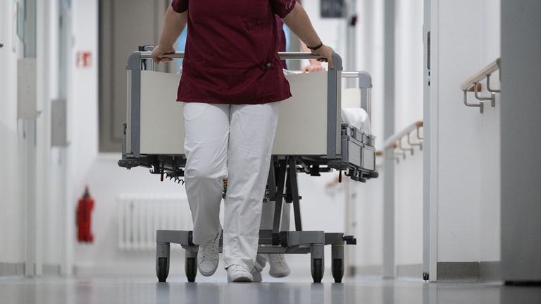 Eine Krankenpflegerin und ein Krankenpfleger schieben ein Krankenbett durch den Gang einer Station eines Krankenhauses.