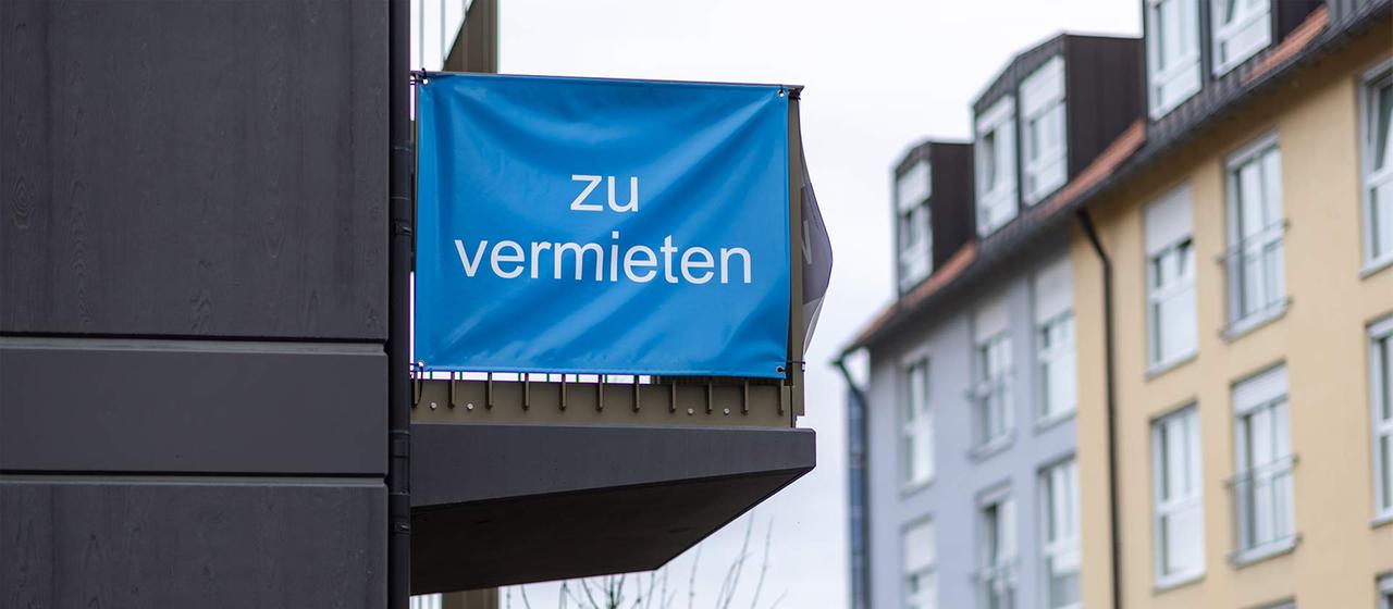 "Zu vermieten" steht auf einer Plane, die an einem Balkon-Geländer einer Neubau-Wohnung angebracht ist.