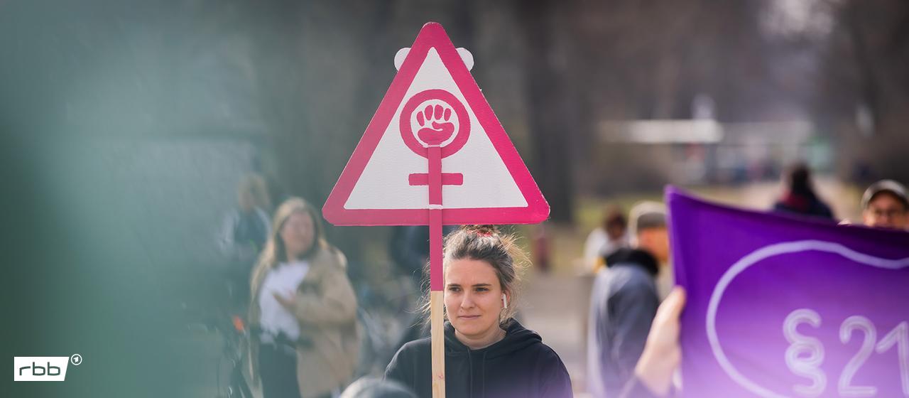 Eine Frau nimmt am 08.03.2026 mit einem Schild mit dem Symbol für Frauen an einer Gewerkschaftsdemonstration unter dem Motto "feministisch, solidarisch, gewerkschaftlich" anlässlich des Internationalen Frauentags teil. (Quelle: Picture Alliance/Christoph Soeder)