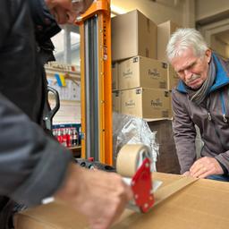 Ein Karton wird von zwei Männern mit einer Paketbandrolle zugeklebt. Im Hintergund Kartons mit der Aufschrift "Pampers". Ganz hinten eine kleine Ukraine-Flagge.