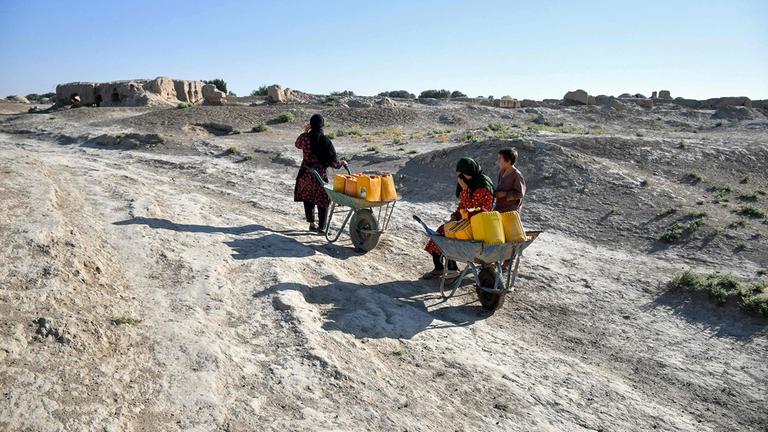 Eine afghanische Frau mit Kindern macht eine Pause, während sie Wasserkanister auf Schubkarren durch eine verlassene Straße im von Dürre heimgesuchten Dorf Bolak im Bezirk Chahar Bolak in der Provinz Balkh am 10. Juli 2025 transportieren. 