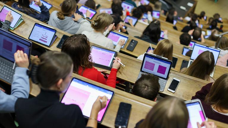 Studierende sitzen in einem Hörsaal einer Universität und schreiben auf ihren Tablets und Laptops