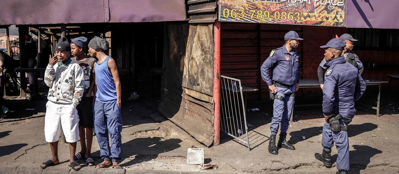 Mehrere Polizisten der South African Police Service stehen vor einem beschädigten Gebäude in Somerset West und beobachten die Umgebung, während einige Männer am Straßenrand stehen und sich unterhalten.