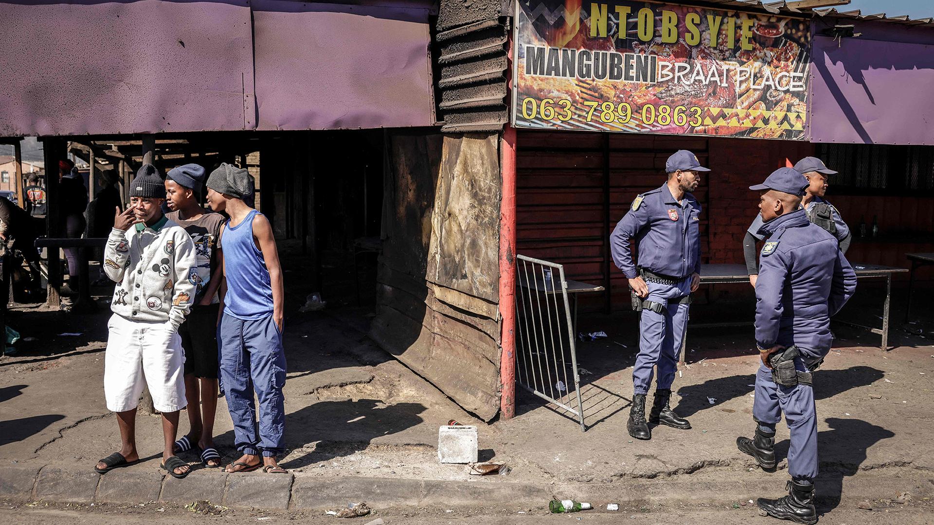 Mehrere Polizisten der South African Police Service stehen vor einem beschädigten Gebäude in Somerset West und beobachten die Umgebung, während einige Männer am Straßenrand stehen und sich unterhalten. | AFP