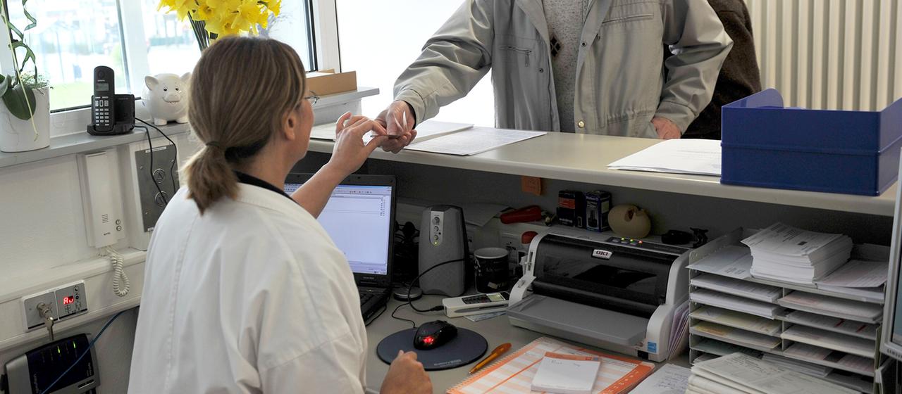 Ein Mann reicht in einer Arztpraxis seine Versichertenkarte über den Tresen.