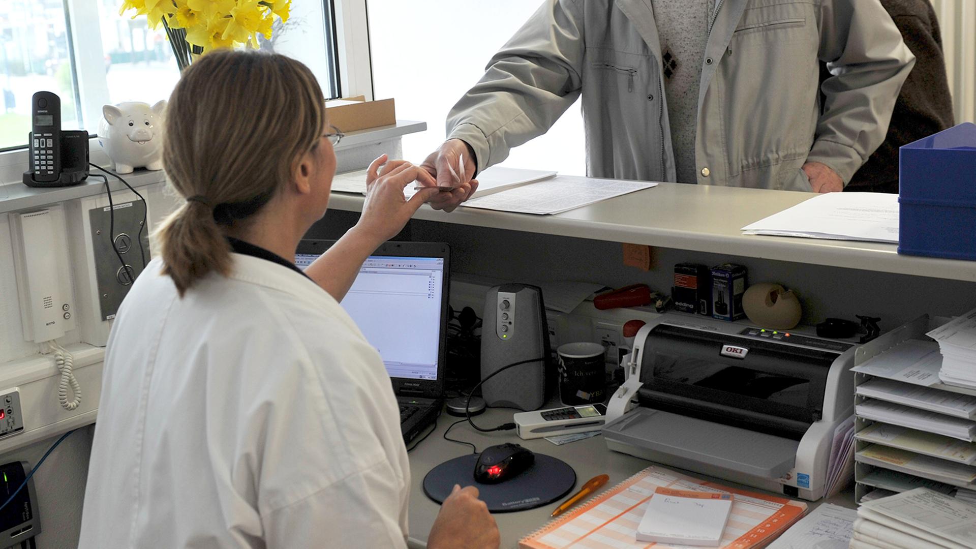 Ein Mann reicht in einer Arztpraxis seine Versichertenkarte �ber den Tresen. | picture alliance / Klaus Rose