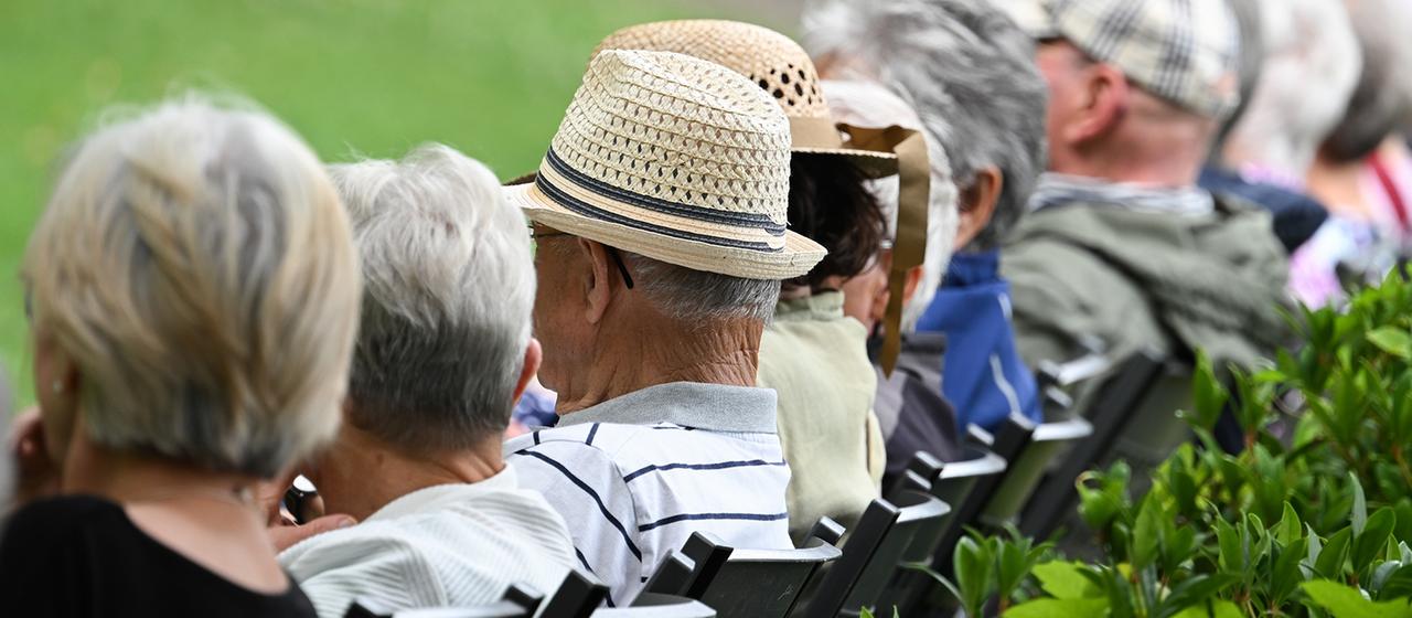 Mehrere ältere Personen sitzen nebeneinander auf Stühlen im Freien.