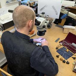 Ein Mann sitzt an einem Tisch und blickt durch ein Mikroskop. Seine Hände arbeiten an einem kleinen Stück, viele kleine Werkzeuge liegen auf dem Tisch. Eine Skizze hängt vor ihm.