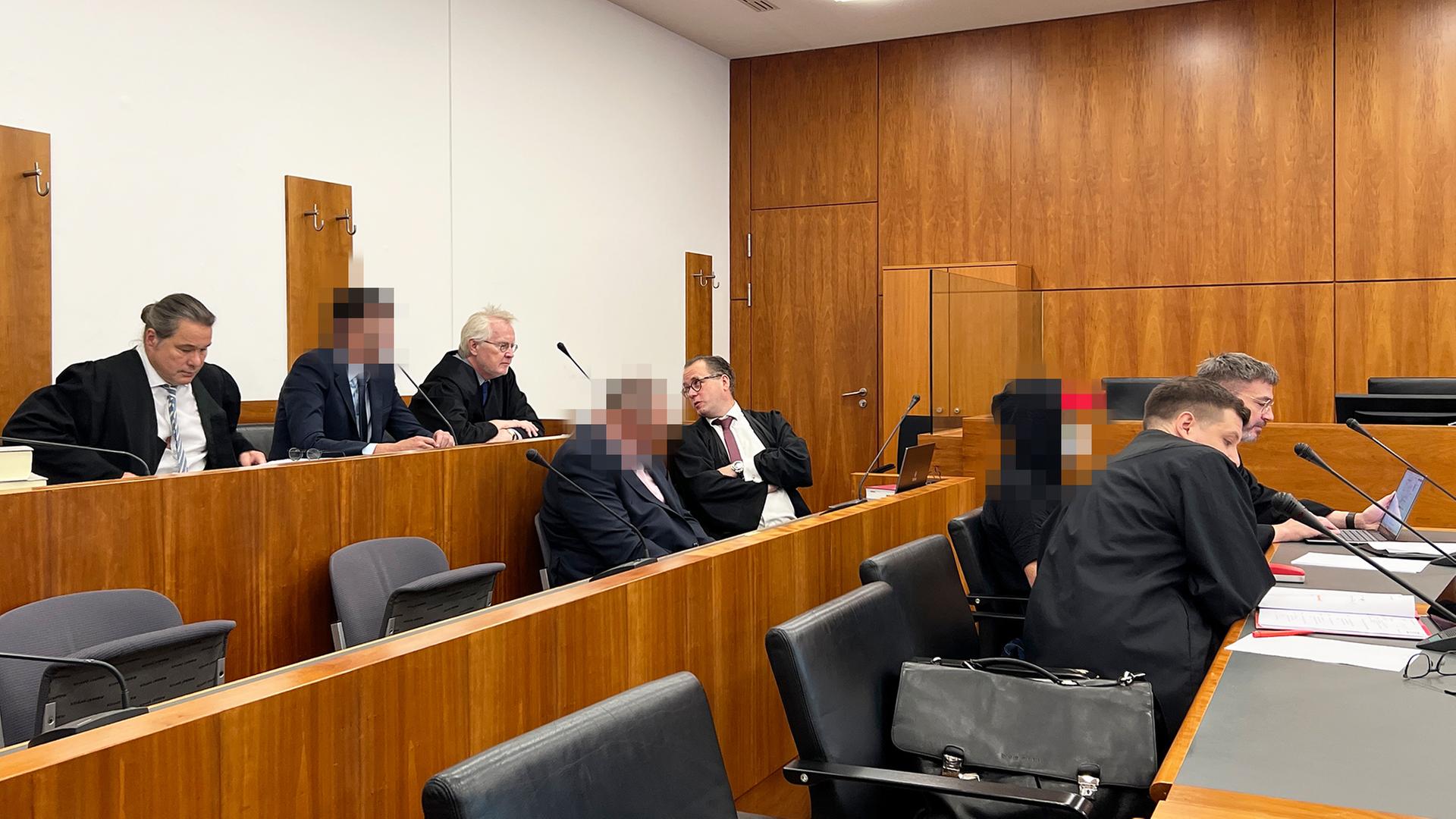 In einem modernen Gerichtssaal mit hözernen Möbeln sitzen drei verpixelte Angeklagte und ihre Verteidiger. | Stefanie Küster (hr)