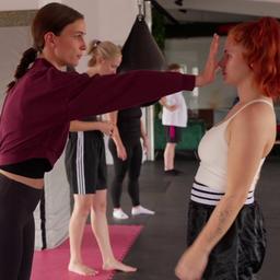 EineTeilnehmerin in einem Selbstverteidigungskurs hält einer anderen Teilnehmerin eine Hand direkt vor das Gesicht.