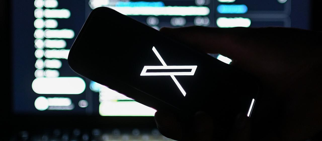Das Logo des Kurznachrichtendiensts X ist auf einem Smartphone zu sehen, das vor die Webversion von X gehalten wird. 