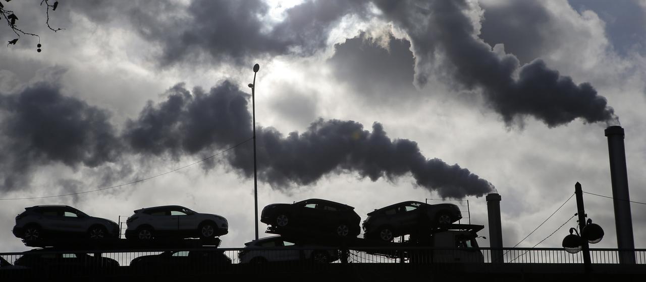 Vor einer Fabrik mit qualmenden Schornsteinen steht ein Zug mit Pkw.