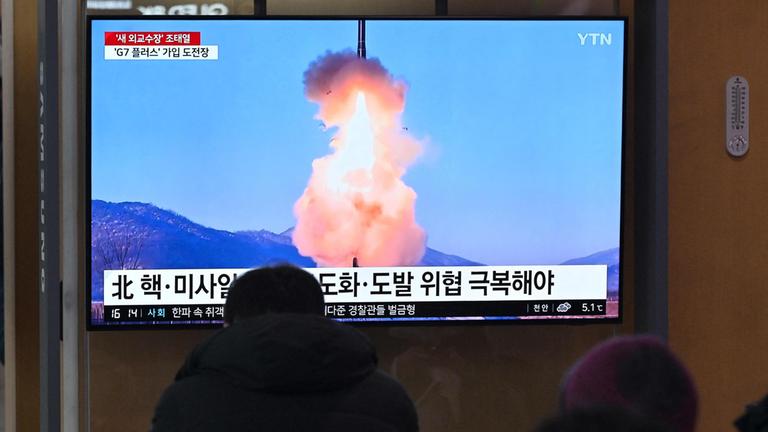 Mehrere Menschen verfolgen den Raketenstart vor einem TV-Bildschirm.