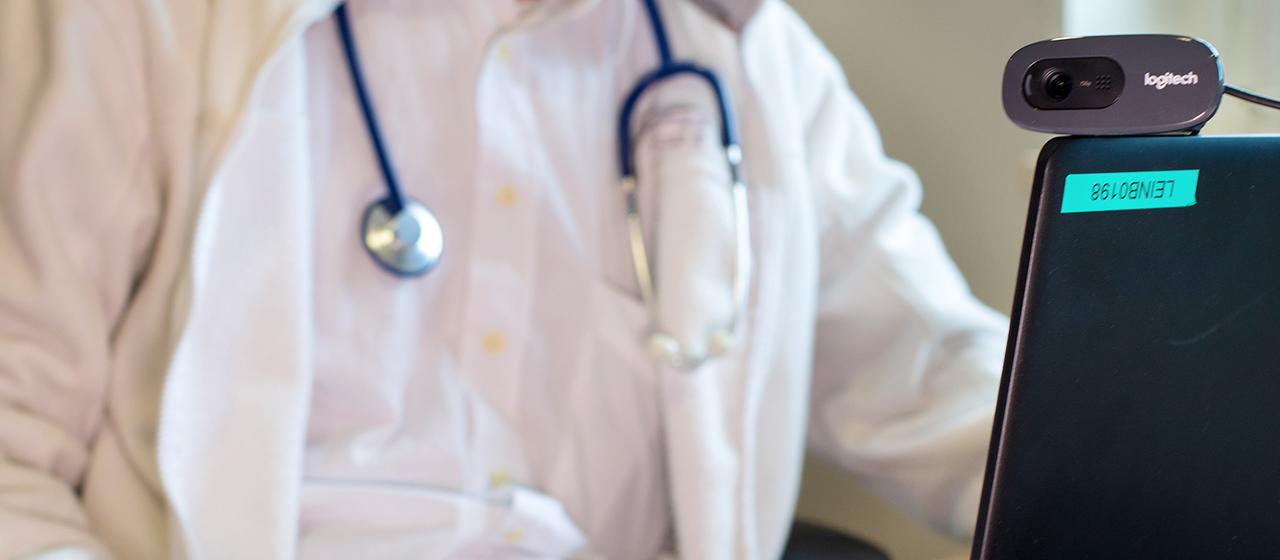 Ein Hausarzt sitzt während einer Videosprechstunde in seiner Praxis vor einem Laptop.