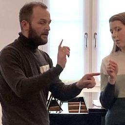 Ein Mann gestikuliert, er scheint zu dirigieren. Eine Jugendliche beobachtet ihn dabei.