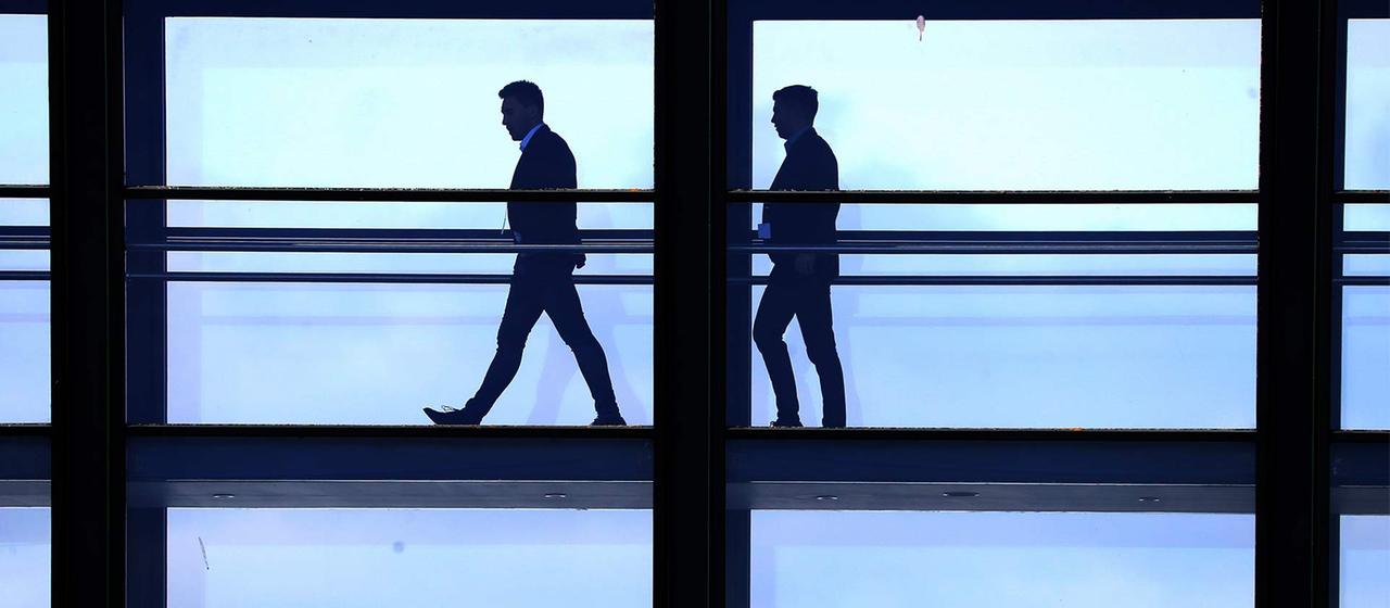 Silhouetten von zwei Männern auf dem Weg durch einen verglasten Gebäudegang.