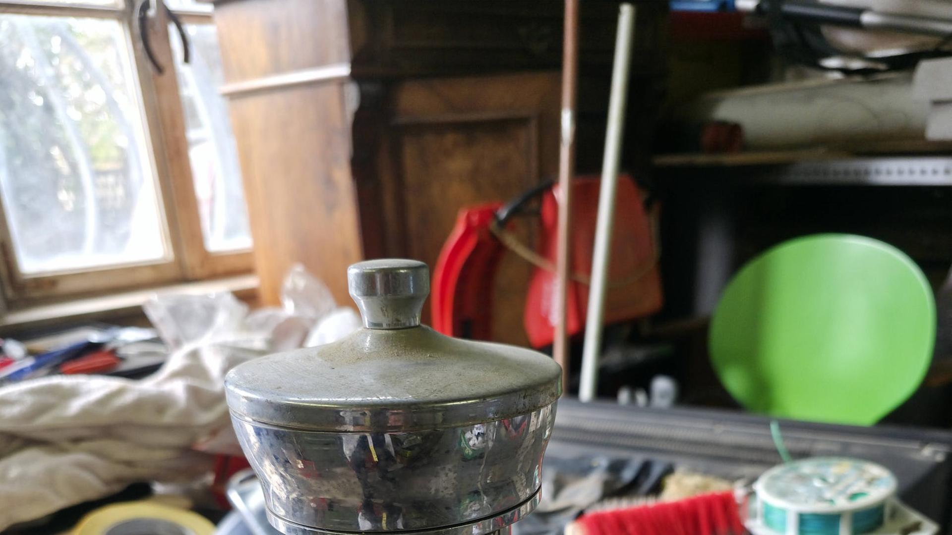 Metallischer Trinkbecher mit Zapfhahn auf einer Werkbank in einer Werkstatt oder einem Lagerraum. Um den Becher liegen verschiedene Werkzeuge und Bürsten. Auf dem Becher steht: „RADIUM TRINK- UND BADESPENDER APPARAT“ | RPDA