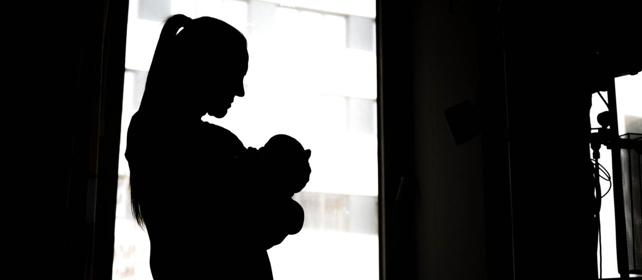 Eine Frau steht mit einem Neugeborenen am Fenster einer Klinik.