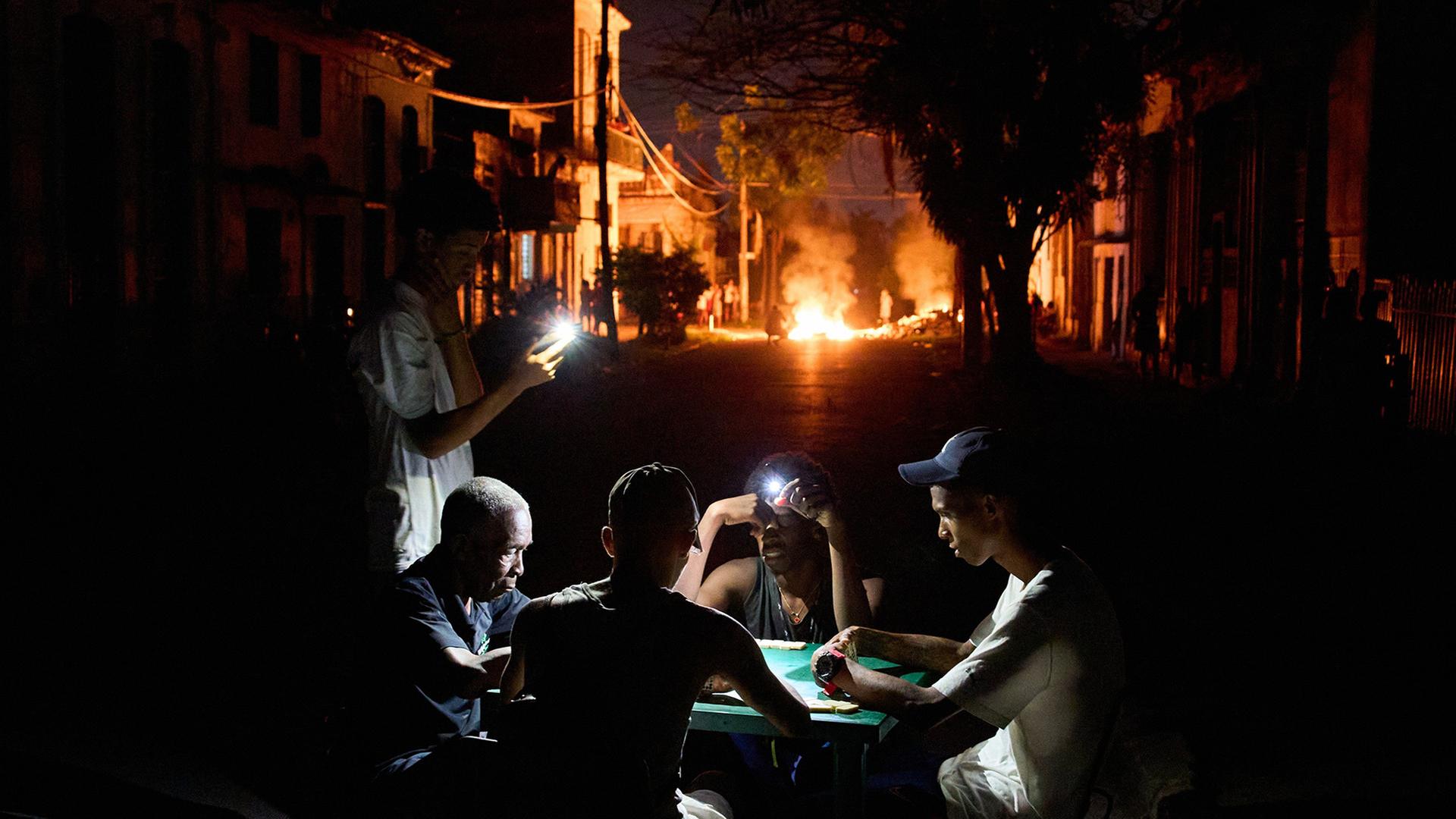 Menschen in der kubanischen Haupstadt Havanna leuchten mit ihren Handys beim Dominospiel, während auf einer Straße ein Feuer brennt. | Ramon Espinosa/AP/dpa