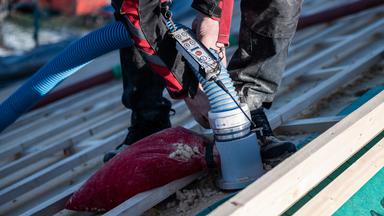Ein Mann schließt am Dach eines Neubaus ein Rohr an, über das Wärmedämmstoff durch eine hochleistungsfähige Einblasmaschine direkt unter das Dach gelangen kann.