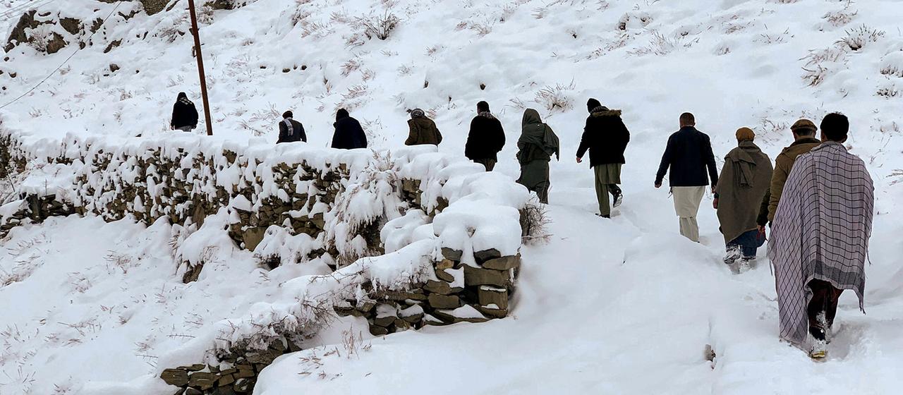 Mehrere Personen laufen auf einem verschneiten Pfad an einer Steinmauer und einem Haus entlang einen Hügel hinauf. 
