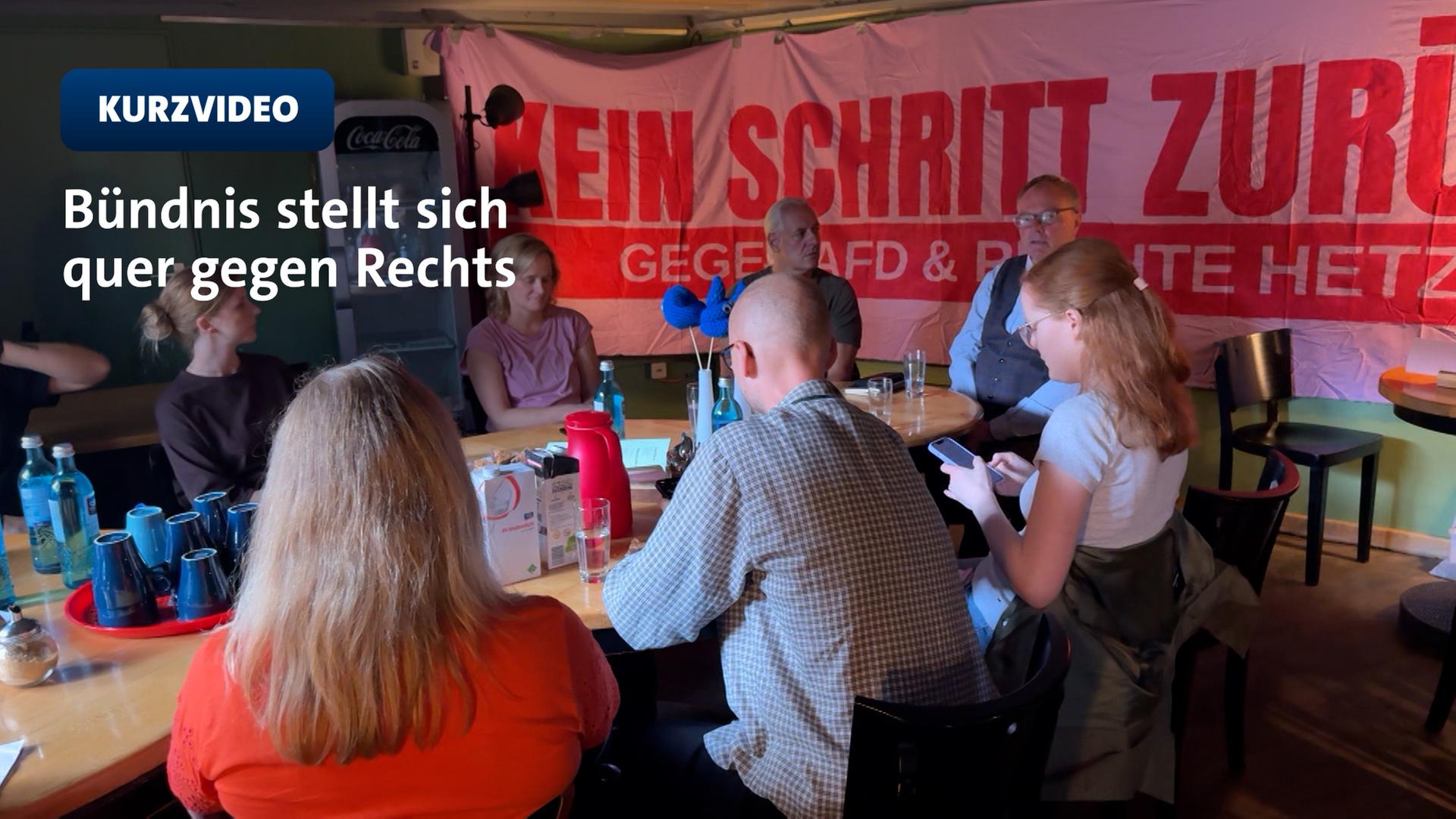 Männer und Frauen sitzen zusammen am Tisch hinter ihnen ein Plakat 