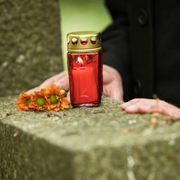 Ein Mann steht an einem Grabstein auf dem sich Blumen und eine Grabkerze befinden.