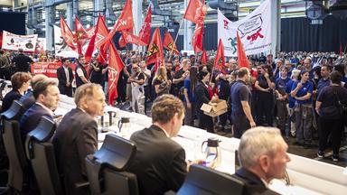 VW-Vorstandsmitglieder sitzen an einem langen schmalen Tisch, vor ihnen demonstrieren Beschäftigte