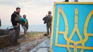 Selenskyj mit blaugelben Blumenstrauß , im Vordergrund ein blaugelbes Schild