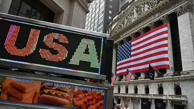 Eine amerikanische Flagge hängt auf der Fassade der New Yorker Börse. Links im Vordergrund der Leuchtschriftzug "USA" auf einem Imbisswagen.